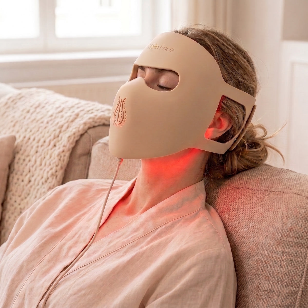 Woman relaxing on a sofa wearing the beige Hello Face M300 silicone LED mask with soft red light