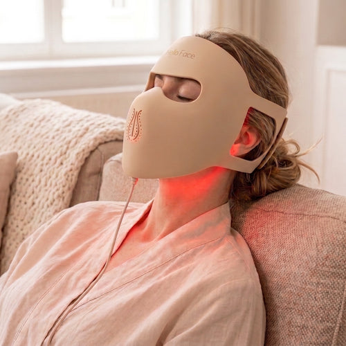 Woman relaxing on a sofa wearing the beige Hello Face M300 silicone LED mask with soft red light
